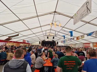 Ein großes Festzelt, gefüllt mit vielen Menschen, die an Tischen sitzen. Im Hintergrund ist eine Bühne zu sehen, auf der eine Veranstaltung stattfindet.
