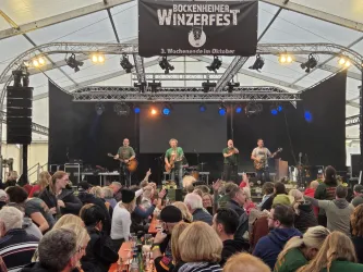 Ein lebhaftes Weinfest mit einer Band auf der Bühne. Viele fröhliche Menschen sitzen an Tischen und genießen ihre Zeit.