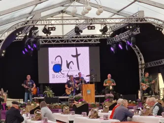 Eine Band spielt auf einer Bühne in einem Festzelt. Im Vordergrund sind Tische mit Besuchern, die das Konzert genießen.