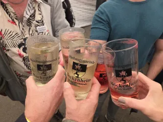 Eine Gruppe von Menschen hebt Gläser mit Getränken an. Die Gläser sind mit dem Wappen von Bockenheim verziert.