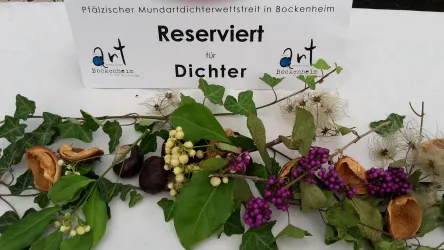 Ein Tisch mit einer Reservierung für Dichter und einer Vielzahl von Kräutern und Blumen dekoriert. Im Hintergrund steht ein Schild mit dem Hinweis auf einen Mundartwettbewerb in Bockenheim.