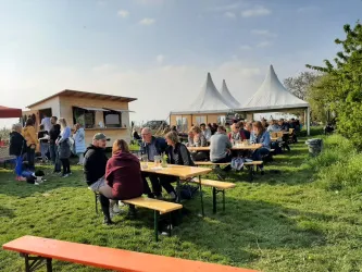 Eine belebte Freiluftveranstaltung mit vielen Menschen an Tischen im Grünen. Im Hintergrund befinden sich ein Verkaufsstand und Zelte.