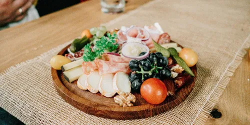 Ein rustikaler Holzuntersetzer mit einer bunten Auswahl an Käse, Wurst, Trauben, Nüssen und frischem Gemüse. Ideal für einen geselligen Abend oder einen Snack mit Freunden.