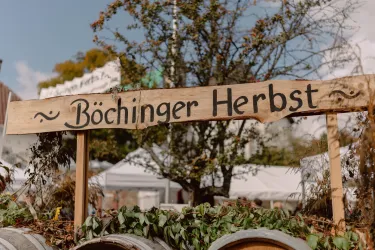 Ein Holzschild mit der Aufschrift "Böchinger Herbst" steht im Vordergrund. Im Hintergrund sind Bäume und eventuelle Marktstände zu sehen.