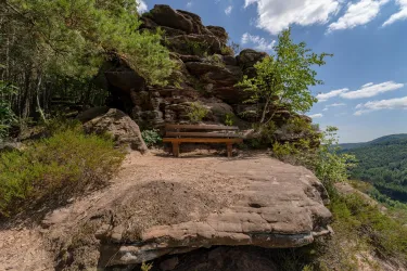 Eine Holzbank auf einem Felsen, umgeben von Bäumen und Sträuchern. Der Himmel ist blau mit einigen Wolken.