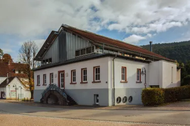 Ein modernes Gebäude mit einem auffälligen Dach und großen Fenstern. Es steht an einer ruhigen Straße, umgeben von Bäumen und Sträuchern.