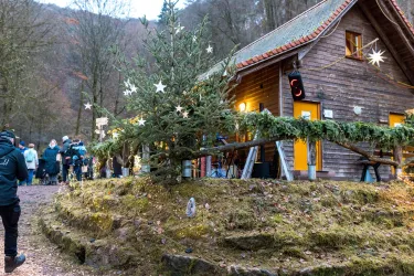 Ein rustikales Holzhaus umgeben von einer malerischen Landschaft. Vor dem Haus steht ein geschmückter Weihnachtsbaum mit Sternen und eine fröhliche Menschenmenge versammelt sich.