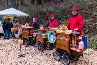 Drei Musikanten in roten Jacken stehen neben nostalgischen Drehorgeln im Wald. Im Hintergrund sieht man Besucher und einen Pavillon.