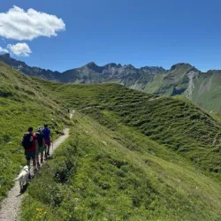 Eine Gruppe von Wanderern geht auf einem schmalen Pfad durch grüne Hügel. Im Hintergrund sind beeindruckende Berge und ein blauer Himmel zu sehen.
