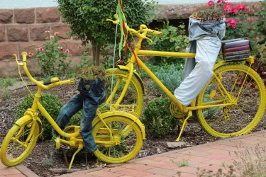 Zwei gelbe Fahrräder mit Blumenkästen, die mit Pflanzen gefüllt sind. Eines der Fahrräder trägt eine Hose als Dekoration.