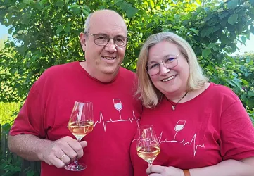 Ein Paar steht vor einem grünen Hintergrund und hält Weingläser in der Hand. Beide tragen rote T-Shirts mit einem Herzschlag-Motiv.