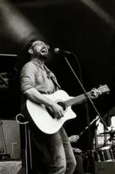 Ein Sänger spielt Gitarre auf einer Bühne. Er trägt eine Brille und hat eine leidenschaftliche Ausstrahlung.