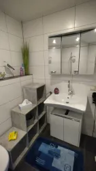 Ein modernes Badezimmer mit einem Waschbecken, einem Spiegel und einer kleinen Ablage. Die Wände sind hell gefliest, und ein blauer Teppich liegt auf dem Boden.