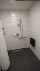Eine moderne Dusche mit weißen Fliesen und einem grauen Boden. Die Dusche ist mit einer Handbrause und einem Duschvorhang ausgestattet.