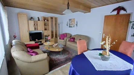 Ein gemütliches Wohnzimmer mit beigen Sofas und pinken Kissen. Ein runder Tisch mit einem weißen Tischläufer und einer Blumendekoration ist ebenfalls zu sehen.