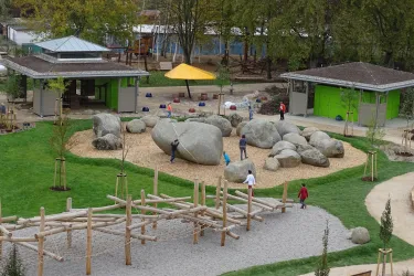 Ein Spielplatz mit großen Steinen und Holzspielgeräten. Kinder spielen und erkunden die Umgebung.