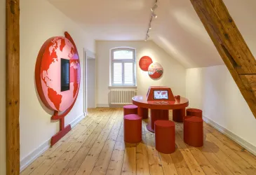 Ein moderner Raum mit einem runden Tisch und Stühlen in Rot. An der Wand befindet sich ein globusförmiges Element sowie ein Bildschirm.