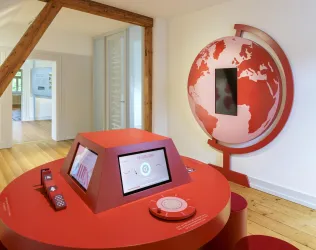 Ein interaktives Ausstellungselement in einem modernen Raum. Es zeigt einen roten Globus und einen runden Tisch mit Bildschirmen.