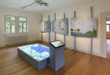 Ein moderner Ausstellungsraum mit Informationsständen an den Wänden. In der Mitte befindet sich ein interaktiver Tisch mit einer Kartenprojektion.