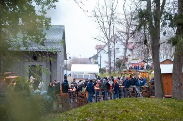 Eine belebte Veranstaltung im Freien mit vielen Menschen, die an Tischen sitzen. Umgeben von Bäumen und kleinen Hütten, strahlt die Atmosphäre festliche Stimmung aus.