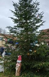 Ein festlich geschmückter Weihnachtsbaum mit Lichtern und einer Holzfigur eines Weihnachtsmanns. Im Hintergrund sind bunte Dekorationen sichtbar.