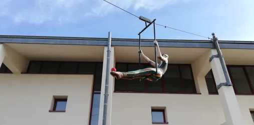 Eine Person schwingt an einem Hochseil und zeigt akrobatische Bewegungen. Der Hintergrund besteht aus einem Gebäude mit großen Fenstern und blauem Himmel.