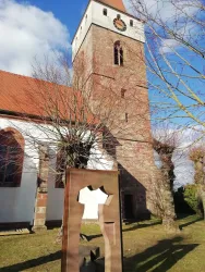 Eine Kirche mit einem hohen Turm und einem kunstvollen Metallkunstwerk im Vordergrund. Der Himmel ist blau mit einigen Wolken, und die Bäume sind noch ohne Blätter.