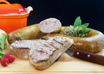 Eine köstliche Wurst in Scheiben geschnitten, umgeben von frischen Kräutern und Gemüse. Im Hintergrund ist ein roter Topf zu sehen.