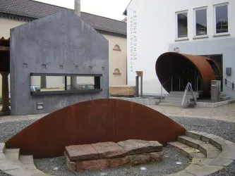Ein moderner Platz mit verschiedenen architektonischen Elementen. Im Vordergrund befindet sich eine runde, metallische Skulptur und im Hintergrund sieht man zwei Gebäude mit unterschiedlichen Baumaterialien.