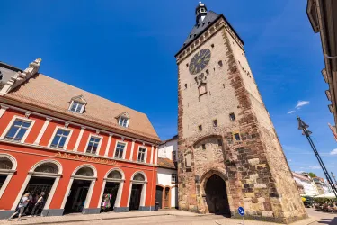 Ein historischer Wasserturm steht neben einem roten Gebäude. Der Himmel ist klar und blau, was die Szene lebendig wirken lässt.