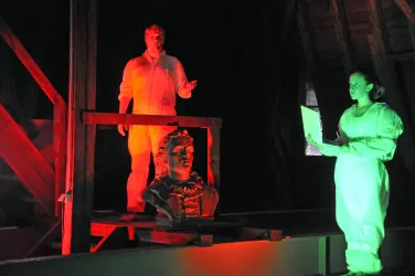 Zwei Personen in Schutzanzügen stehen in einem beleuchteten Raum. Eine Person liest von einem Blatt, während die andere spricht und eine Statue im Hintergrund beleuchtet ist.