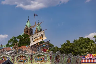 Ein historisches Gebäude mit Türmen und einem Segel auf einem Schiff. Darunter befindet sich ein bunter Freizeitpark mit Schildern und dekorativen Elementen.