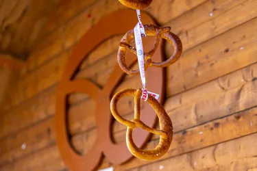 Eine traditionelle Brezel hängt an einer Holzwand. Im Hintergrund ist ein stilisiertes Brezel-Symbol zu sehen.