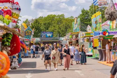 Ein belebter Jahrmarkt mit bunten Ständen und vielen Besuchern. Im Hintergrund sind grüne Bäume und ein blauer Himmel zu sehen.