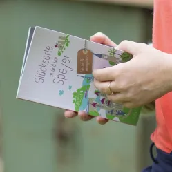 Eine Person hält ein Buch mit dem Titel "Glücksorte in und um Speyer". Das Buch zeigt bunte Illustrationen und scheint Informationen über interessante Orte zu bieten.