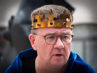 Ein Mann trägt eine goldene Krone mit roten Edelsteinen. Er hat einen ernsten Gesichtsausdruck und trägt einen blauen Umhang.