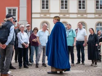 Eine Gruppe von Touristen hört einem Führer in einem blauen Umhang und einer Krone zu. Die Szene spielt sich auf einem Platz mit historischen Gebäuden im Hintergrund ab.