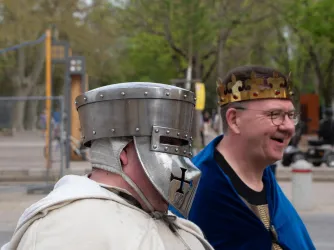 Zwei Männer in historischen Kostümen stehen nebeneinander. Einer trägt einen Metallhelm, der andere eine Krone und einen blauen Umhang.