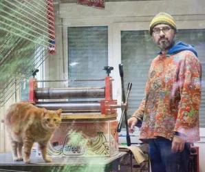 Ein Mann steht in einem kreativen Atelier neben einer orangefarbenen Katze. Im Hintergrund sieht man eine Druckmaschine und einen hellen Raum.