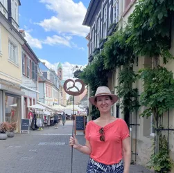 Eine Frau mit einem Hut hält ein Schild in Form einer Brezel in einer belebten Straße. Im Hintergrund sind Cafés und Geschäfte sowie ein wolkenloser Himmel zu sehen.