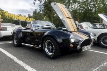 Ein schwarzes Sportauto mit offenen Motorhaube steht auf einem Parkplatz. Im Hintergrund sind weitere Fahrzeuge und Bäume zu sehen.