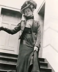 Eine elegant gekleidete Frau steht auf einer Treppe. Sie trägt einen auffälligen Hut und hat einen selbstbewussten Ausdruck.