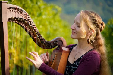 Eine Frau spielt Harfe vor einer natürlichen grünen Kulisse. Sie lächelt und genießt die Musik.