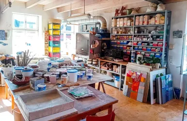 Ein kreatives Atelier mit Tischen voller Malutensilien und Farbtöpfen. Regale mit verschiedenen Kunstmaterialien und Farbpaletten füllen den Raum.