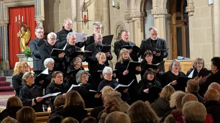 Ein Chor steht in einer Kirche und singt. Die Mitglieder tragen schwarze Kleidung und haben Notenblätter in der Hand.