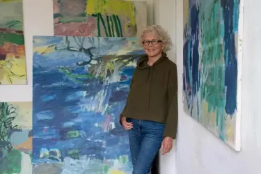 Eine Frau steht in einem Kunststudio und lächelt. Im Hintergrund sind mehrere farbenfrohe Gemälde zu sehen.