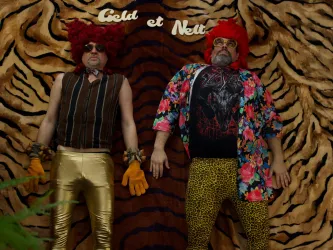 Zwei Männer in auffälligen Kostümen liegen auf einem Tierfell-Hintergrund. Sie tragen bunte Hemden und haben leuchtend rote Perücken auf.