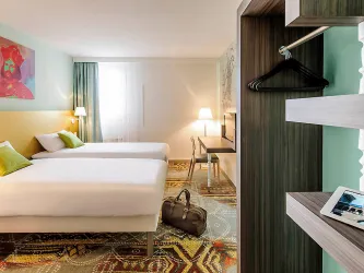 Ein modernes Hotelzimmer mit zwei Einzelbetten, einem Schreibtisch und einem großen Fenster. Die Wände sind in sanften Farben gehalten, und der Boden hat einen bunten Teppich.