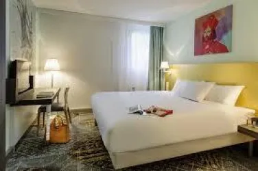 Ein modernes Hotelzimmer mit einem großen Doppelbett und frischer Dekoration. Es gibt einen Schreibtisch, einen Stuhl und angenehmes Licht durch die Fenstervorhänge.