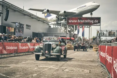 Eine Oldtimer-Rallye findet auf einem Flughafen statt. Im Hintergrund fliegt ein Flugzeug über die Rennstrecke.